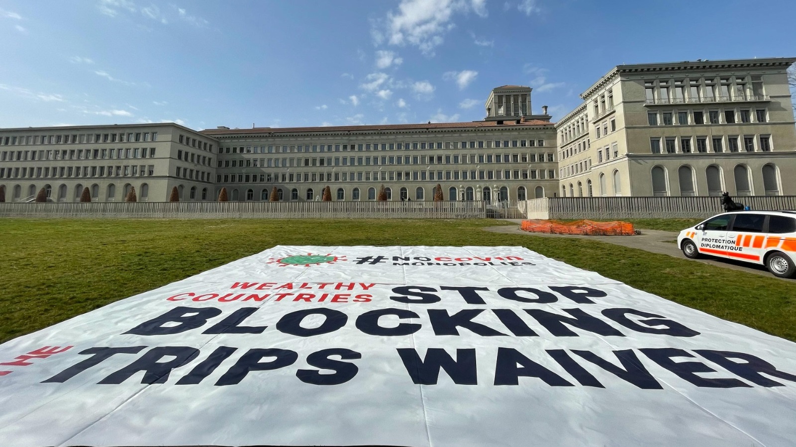 Large cloth banner on the lawn of the World Trade Organization headquarters reading 'Wealthy countries stop blocking TRIPS waiver.'