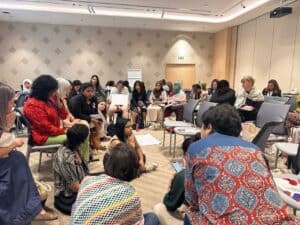 A group discussion happening in a warmly lit room among several women. Many of them are sitting on chairs while a few are on the carpeted floor."