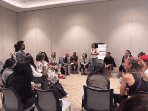 Group of participants, mostly women, having a discussion in warmly lit room. They are sitting on gray-colored chairs arranged in a circle. Two of the participants are standing and having a conversation while others are listening."